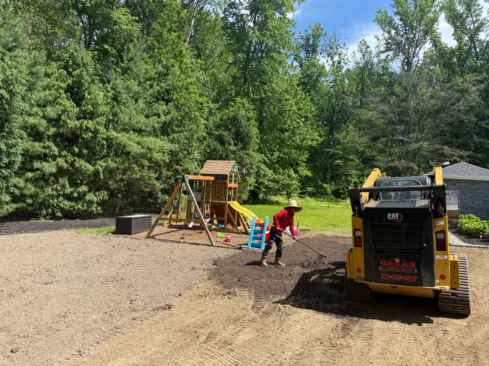 Worker using a skid steer to spread compost in a residential yard with trees and a playground, emphasizing soil amendment and landscaping services by Urban Landscapes in Princeton, NJ.