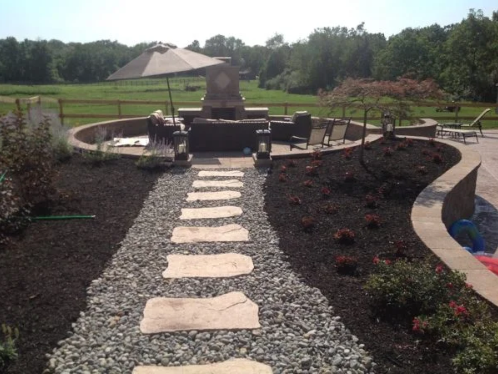 Landscape design featuring a mulched garden path with stone stepping stones, surrounded by flowering plants and a seating area under an umbrella, showcasing proper mulch application for aesthetic and plant health.