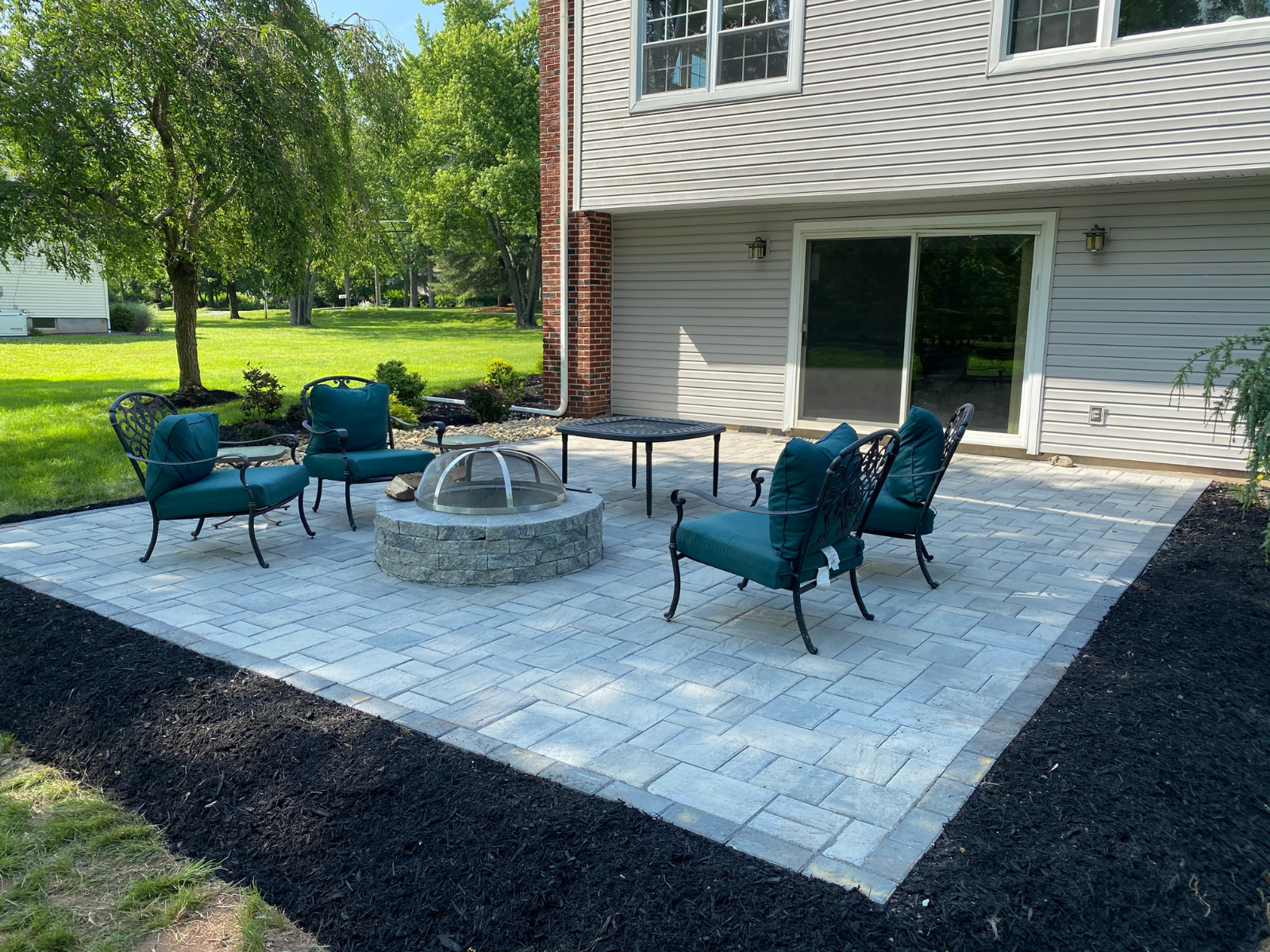 Patio area featuring green cushioned chairs around a stone fire pit, surrounded by landscaped grass and mulch, illustrating outdoor living space design.