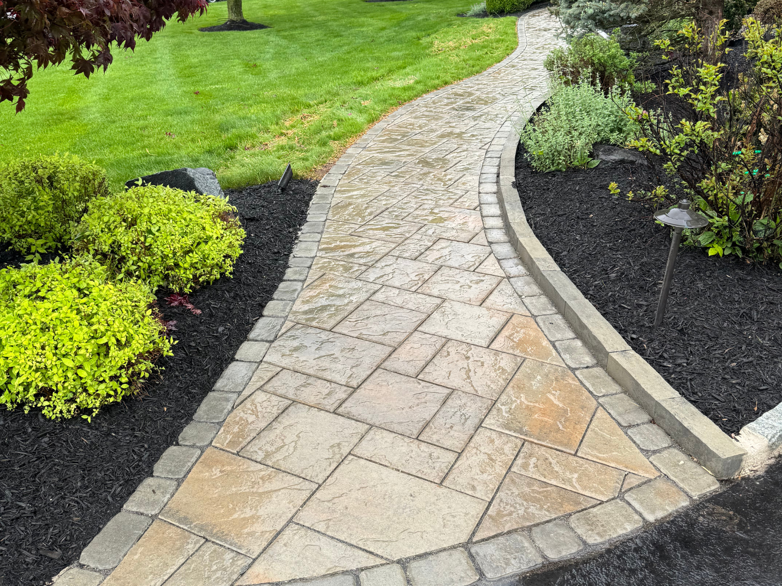 Curved walkway with stone pavers, bordered by lush green shrubs and black mulch, showcasing a well-lit path for safe navigation in landscaping design.