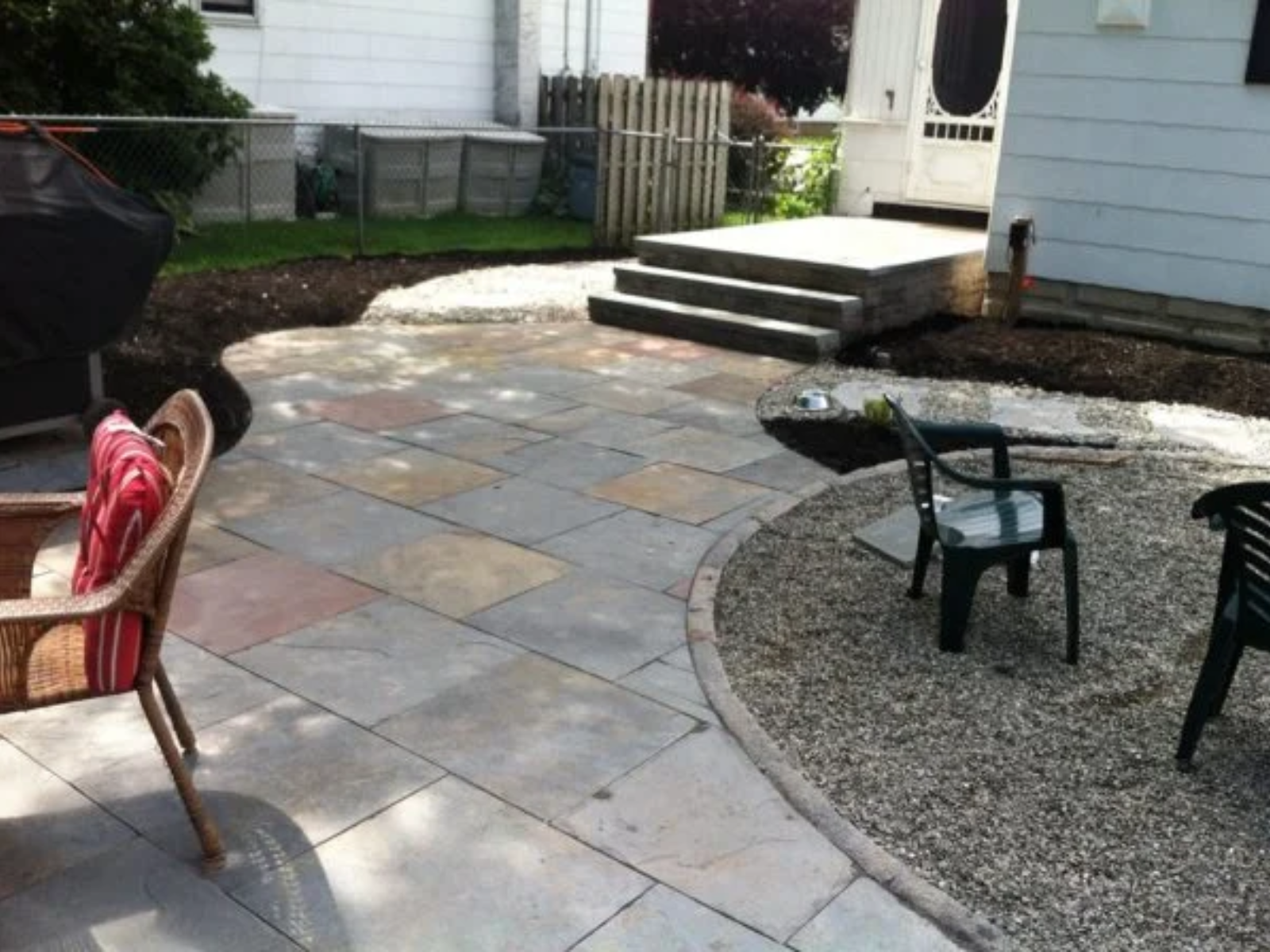 Patio area with stone pavers, wicker chair with red cushion, green plastic chairs, and freshly mulched garden beds in Princeton, NJ, highlighting landscaping services by Urban Landscapes.