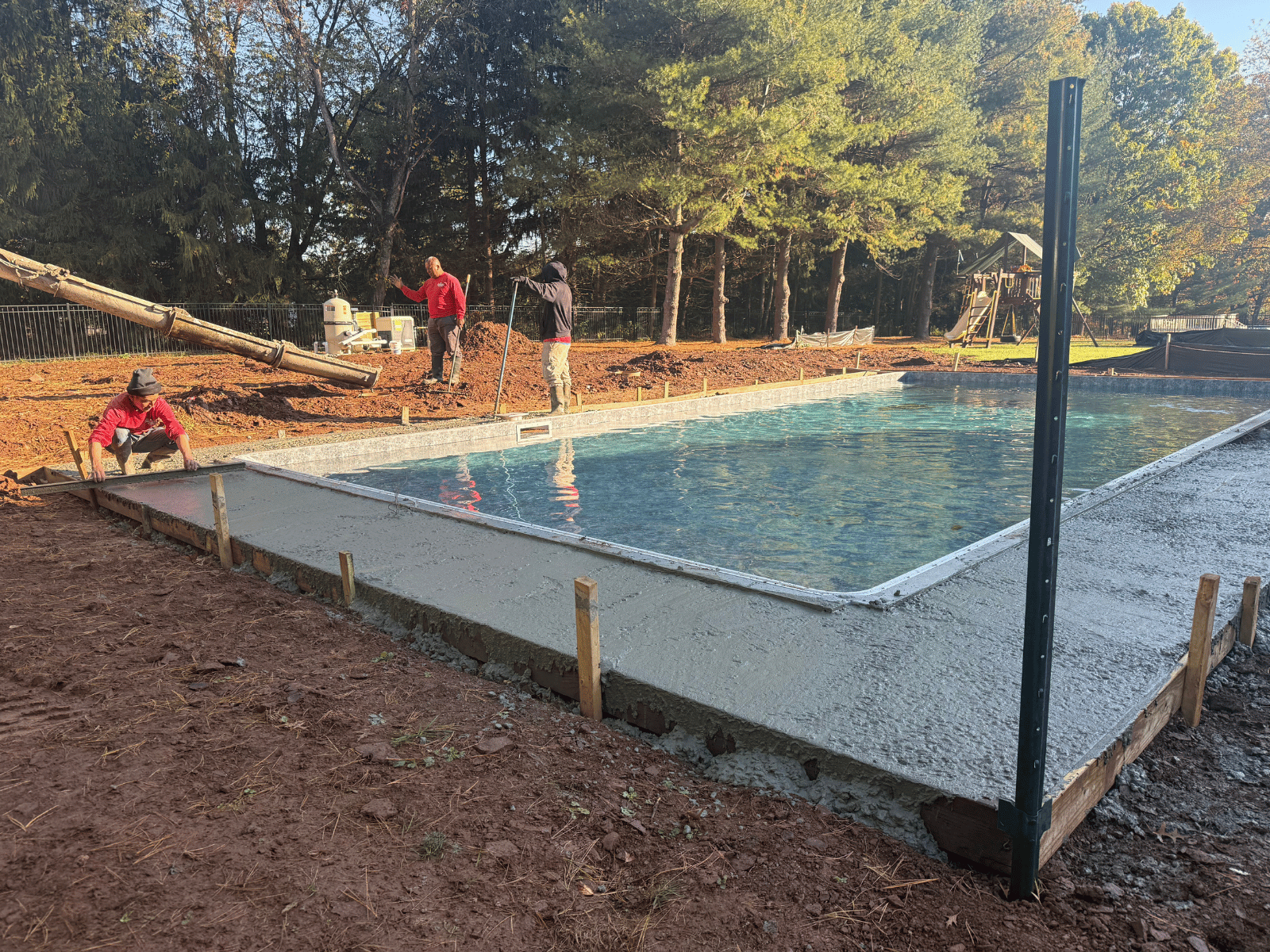 Workers installing a concrete swimming pool deck beside a newly constructed pool in a residential backyard in Princeton, NJ, with trees and landscaping in the background.