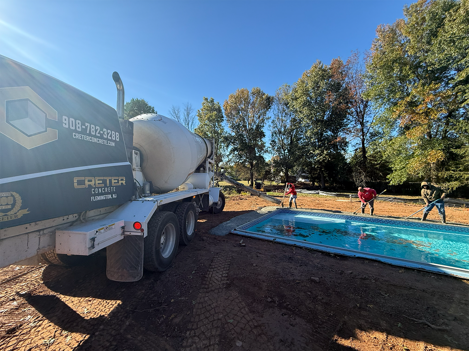 Concrete truck delivering material for hardscaping project, workers preparing pool area with tools, surrounded by trees in residential landscape.