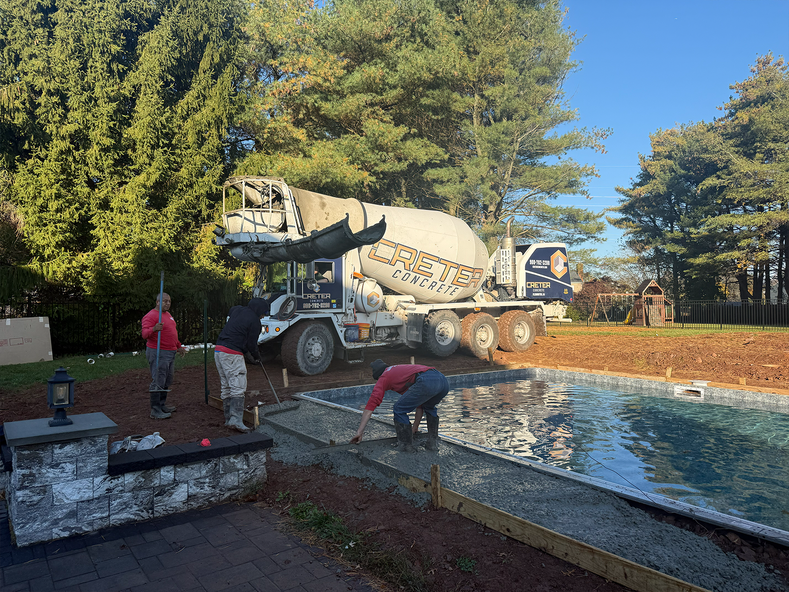 Construction crew pouring concrete for hardscaping installation near a swimming pool, with a concrete mixer truck in the background, showcasing landscaping transformation in a residential outdoor space.