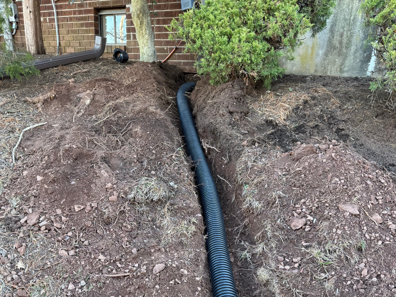 Drainage installation with black corrugated pipe in a trench, surrounded by soil and vegetation near a building, illustrating custom landscape drainage solutions.