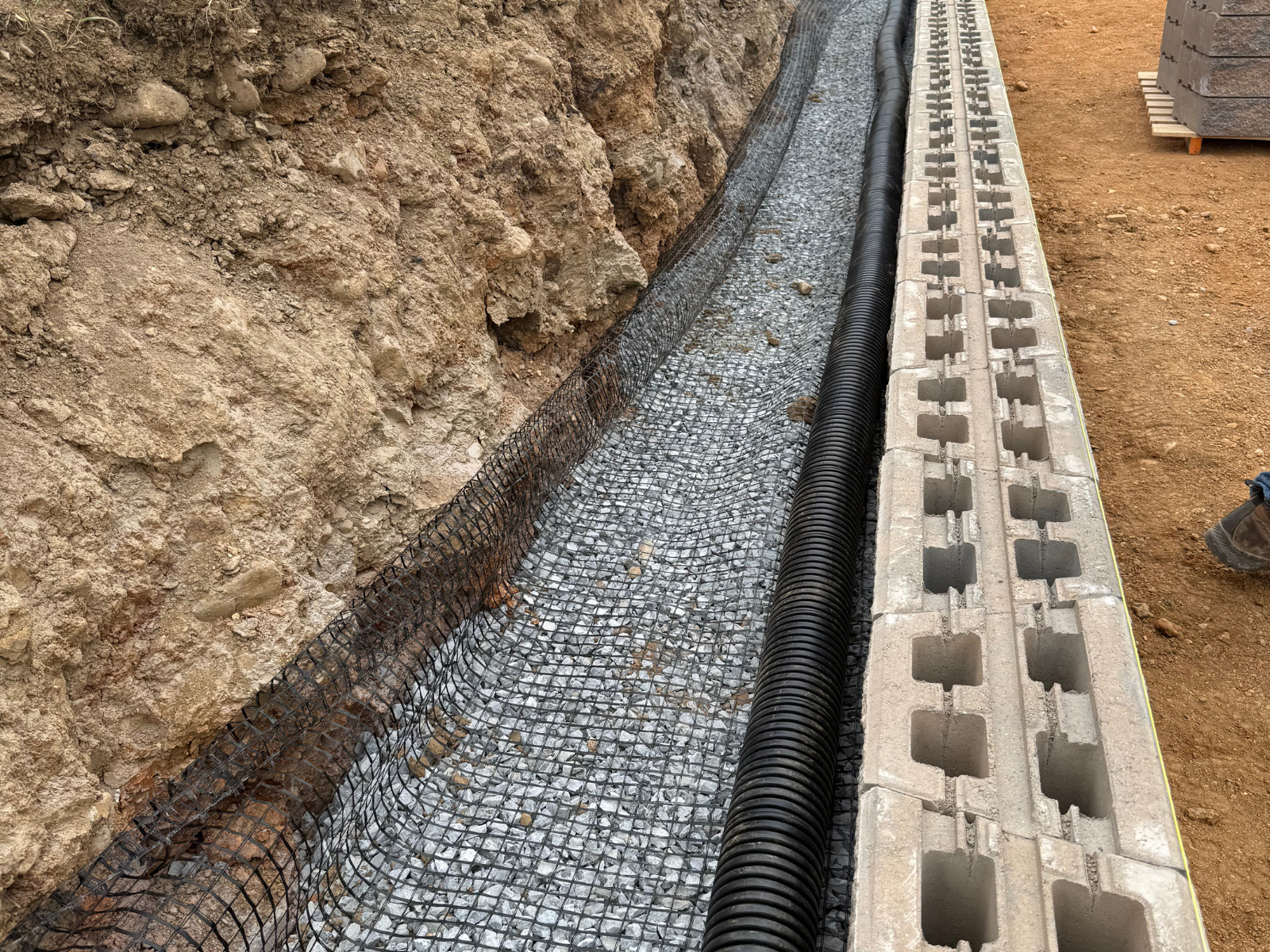 Retaining wall construction site showing gravel base, drainage pipe, and foundation materials, highlighting engineering and material selection for durability and aesthetics in landscaping.